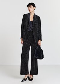 Schwarzer Samtanzug mit weit geschnittenen Hosen und einem Blazer, der einen subtilen Glanz hat. Kombiniert mit einer schwarzen Pelztasche und patentierten High Heels.