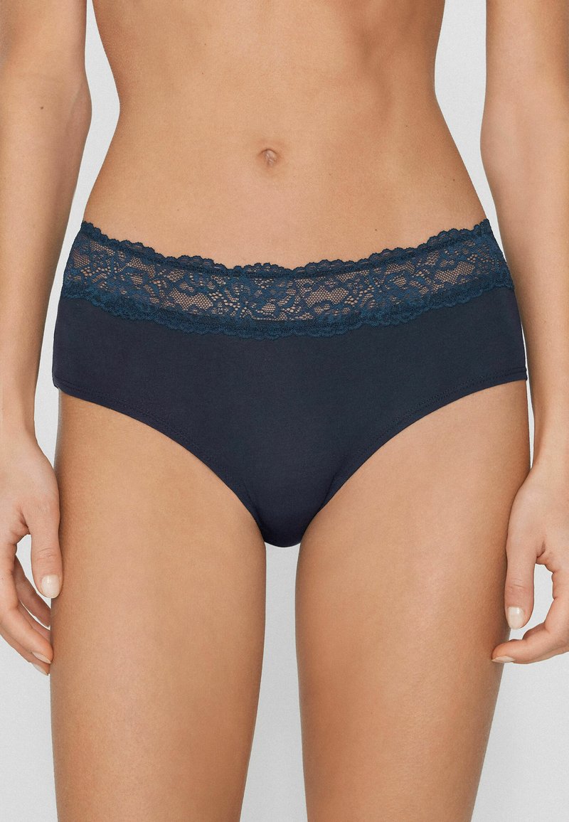 Mujer con braguitas de tiro alto color azul marino con cinturilla ancha de encaje, de pie con los brazos relajados a los lados, contra un fondo liso.