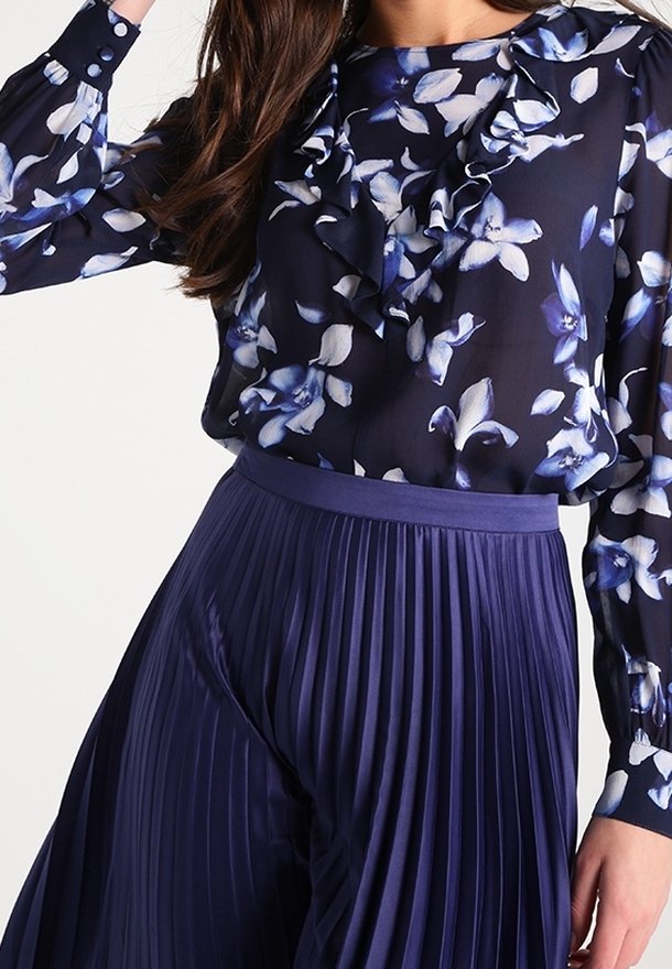 Blusa a stampa floreale con ruches in blu navy, abbinata a pantaloni plissettati blu scuro. Tessuto liscio con una leggera lucentezza e design strutturato.