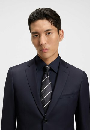 Veste de costume marine avec un revers cranté, assortie à une chemise bleu foncé et une cravate rayée grise. Tissu lisse avec une silhouette ajustée.