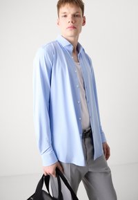 Camisa de manga larga azul claro con botones en el frente, textura suave y un cuello, combinada con pantalones grises. Modelo sosteniendo un bolso negro.
