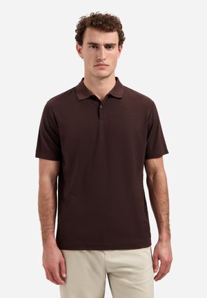 Mann mit lockigem Haar, der ein dunkelbraunes Kurzarm-Poloshirt und beigen Hosen trägt, steht vor einem schlichten hellen Hintergrund.