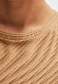 Detailní záběr na krk a horní část hrudníku osoby oblečené v béžovém texturovaném pleteném svetru s kulatým výstřihem.