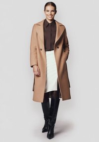 Płaszcz z mieszanki wełny w kolorze camel, z oversized kołnierzami, podwójnymi guzikami, dopasowaną brązową bluzką i białą spódnicą, w zestawieniu z czarnymi kozakami do kolan.