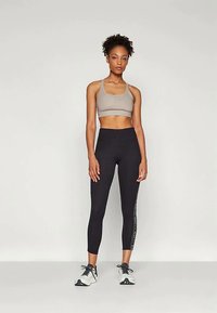 Športová podprsenka v svetlo šedej farbe, nohavice s vysokým pásom v čiernej farbe s lesklým logom na boku, v kombinácii so šedými a bielymi teniskami. Jednoduché pozadie.