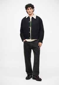 Chaqueta de mezclilla negra con un cuello de borreguillo crema, llevada sobre una camisa blanca y combinada con jeans de pierna ancha de lavado oscuro y zapatos negros.