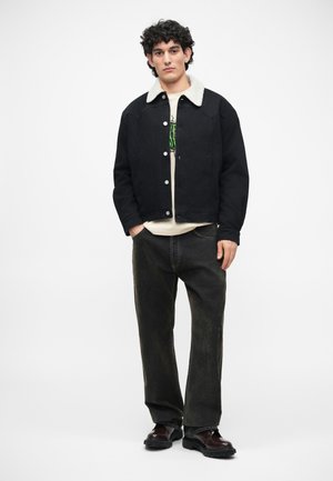Veste en denim noir avec un col en fausse fourrure crème, portée sur une chemise blanche et assortie avec un jean large à délavage foncé et des chaussures noires.