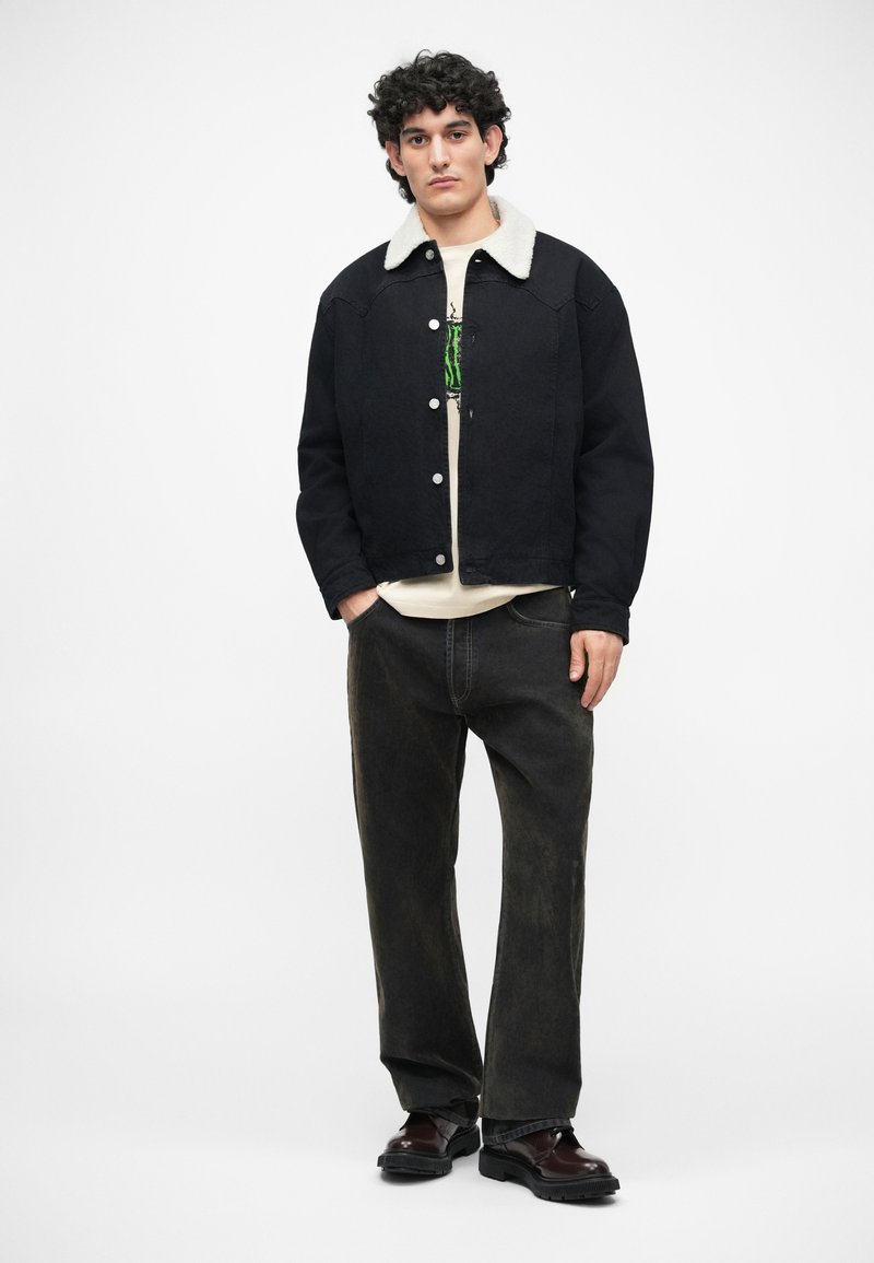 Veste en denim noir avec un col en fausse fourrure crème, portée sur une chemise blanche et assortie avec un jean large à délavage foncé et des chaussures noires.