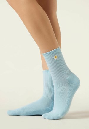 Beine in hellblauen, gerippten Socken mit einer kleinen, eingestickten weißen und gelben Blume an der Außenseite der rechten Socke.