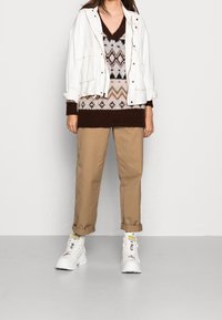 Veste blanche, pull en maille marron à motifs, pantalon beige et baskets blanches épaisses avec des accents jaunes. Silhouette ajustée et décontractée.