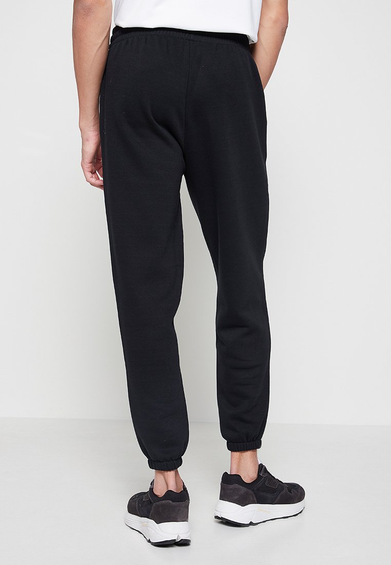 Czarne dresowe spodnie z elastycznym pasem i mankietami u kostek, wykonane z miękkiego materiału. Noszone z czarnymi butami sportowymi. Prosty, funkcjonalny design.