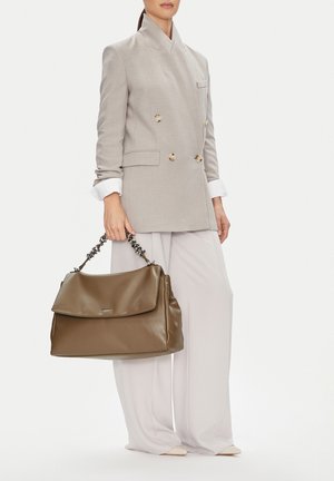 Žena v béžovém dvouřadém saku a bílých širokých kalhotách, držící velkou hnědou koženou kabelku s řetízkovým uchem.