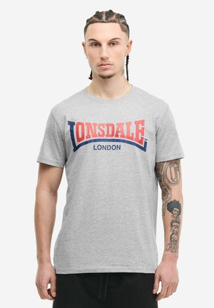 Uomo con capelli intrecciati che indossa una maglietta grigia Lonsdale London e pantaloni neri, mostrando tatuaggi dettagliati sul braccio sinistro.