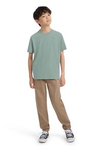 Fant s stoji z rokami sproščenimi, oblečen v zeleno majico, bež hlače in črne superge z belimi vezalkami na belem ozadju.