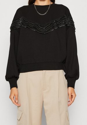 Pull noir avec un ourlet côtelé et des manches ballon, présentant des volants superposés ornés de sequins argentés sur la poitrine.