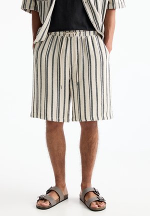 Shorts crème pour hommes arrivant au genou avec des rayures noires verticales, taille à cordon, assortis à une chemise coordonnée et des sandales grises à boucle.