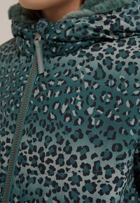 Pūkinė striukė su mėlynosios žalios spalvos pagrindu, dekoruota juodu leopardų raštu. Ji turi kailinį gobtuvą ir užtrauktuką su pilkos spalvos detalėmis.