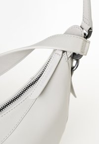 Borsa a mano bianca in pelle con forma curva, dotata di una cerniera nera, cuciture grigie e tracolla regolabile. Texture liscia e dettagli hardware minimi.