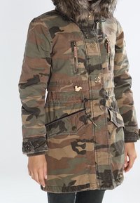 Kamuflażowy zimowy płaszcz z kapturem wykończonym futrem, zamykanymi kieszeniami na piersi, regulowanym sznurkiem w talii oraz zapinanymi kieszeniami na biodrach.
