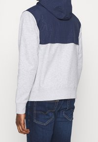 Sudadera gris con sección superior y capucha en color azul marino. La zona azul marino presenta un patrón en relieve. Combinada con jeans azul oscuro.