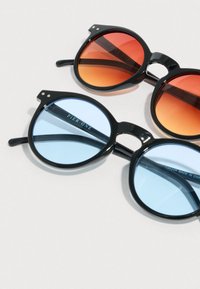 Δύο ζευγάρια στρογγυλά μαύρα γυαλιά ηλίου με φακούς με διαβαθμισμένο χρώμα, το ένα πορτοκαλοκόκκινο και το άλλο ανοιχτό μπλε, τοποθετημένα σε λευκή επιφάνεια.