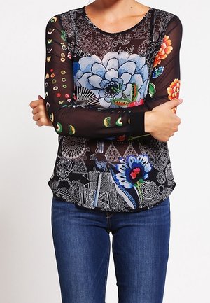 Personne portant un top à manches longues en maille noire avec des motifs floraux et géométriques colorés, associée à un jean bleu, les bras croisés.