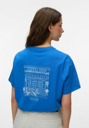 Vrouw met lang bruin haar die een blauw T-shirt draagt met een witte lijntekening van een koffiebar en de tekst "New York East Village" op de achterkant.