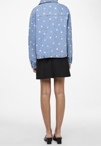 Jeansjakke med gule tusindfryd broderier, lyseblå, med krave, knaplukning ved ærmerne, parret med en sort nederdel og højhælede sandaler.