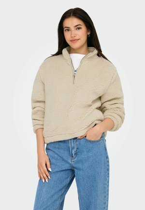 Beige bolyhos pulóver félzippes gallérral, texturált felülettel és leengedett vállakkal, világoskék farmernadrággal párosítva.