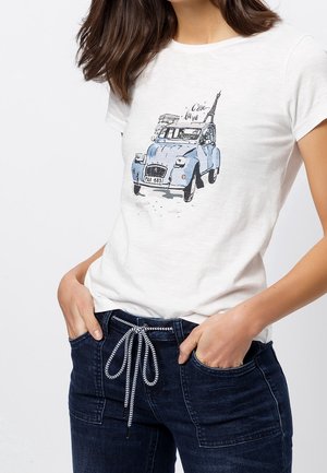 Femme portant un t-shirt blanc avec une illustration d'une voiture vintage bleue et de la tour Eiffel et le texte "C'est la vie" sur le devant, associé à un jean foncé.
