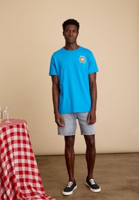 Blauwe katoenen T-shirt met grafische print op de borst, gecombineerd met lichtdenim shorts en marineblauwe sneakers. Eenvoudige, casual uitstraling.