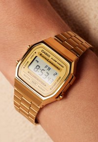 Montre digitale en or avec un cadran carré, bracelet en maillons métalliques et un affichage lumineux montrant l'heure et des fonctions supplémentaires. Résistante à l'eau.