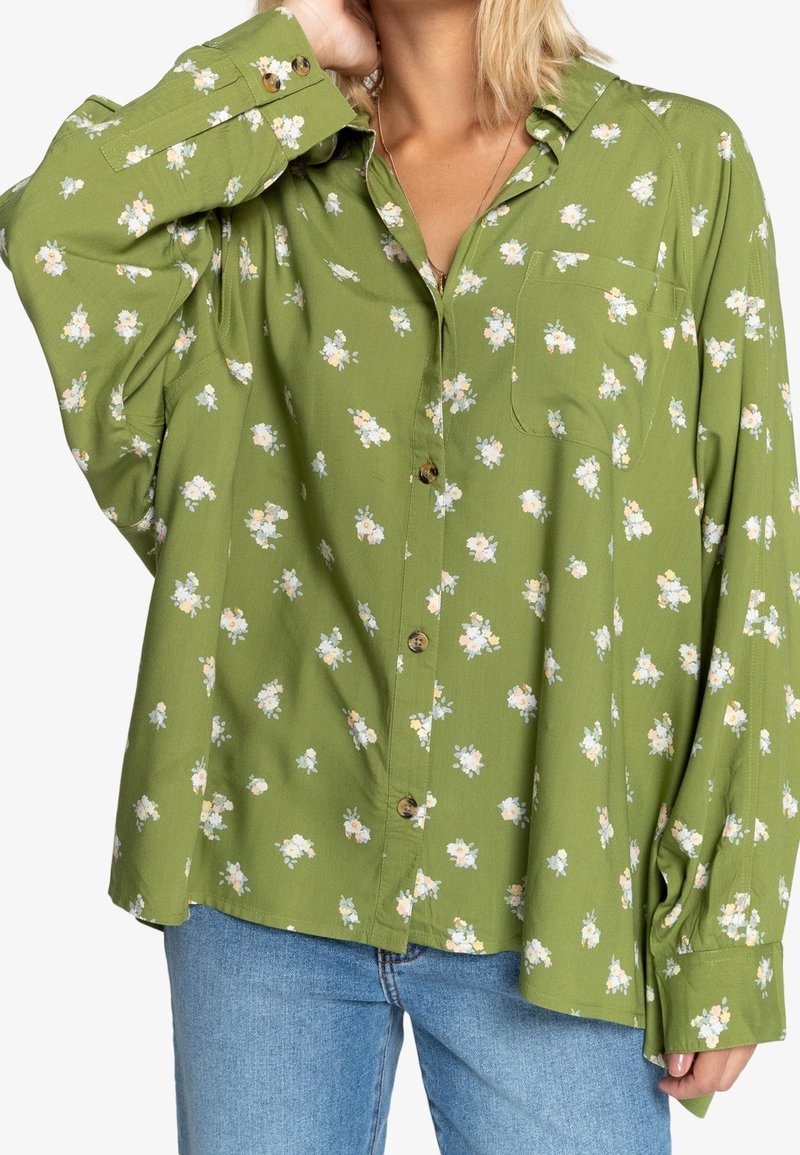 Groene bloemenoverhemden van lichtgewicht stof. Heeft kleine witte en gele bloemen, lange mouwen en een borstzak.