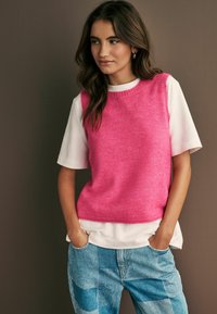 Rosa stickad ärmlös tröja över en vit kortärmad skjorta, parat med blå lapptäcksjeans. Mot en neutral bakgrund.