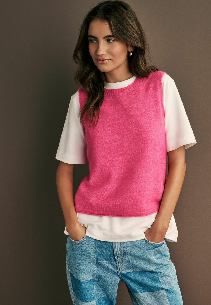 Rosa stickad ärmlös tröja över en vit kortärmad skjorta, parat med blå lapptäcksjeans. Mot en neutral bakgrund.