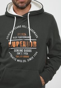 Sudadera gris oscuro con capucha forrada en blanco, que presenta un destacado diseño gráfico en texto naranja y blanco. Incluye un bolsillo tipo canguro en la parte frontal.
