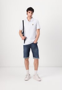 Weißes Poloshirt mit kleinem gelbem Logo, kombiniert mit hochgekrempelten Denim-Shorts und weißen Sneakers. Schlichte, legere Kleidung mit einem marineblauen Armband.