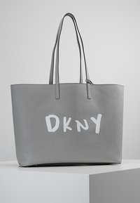 Sac cabas gris avec de longues anses et un logo DKNY blanc audacieux à l'avant, posé sur une surface blanche devant un fond gris.