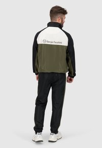 Tuta da uomo in tessuto sportivo di colore nero, verde e bianco; presenta una giacca con zip e logo sul retro, polsini elasticizzati e pantaloni in stile jogger.