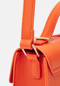 Détail d'un sac à main en cuir orange avec une sangle cousue, une quincaillerie dorée et une silhouette structurée. Texture lisse avec des lignes épurées.