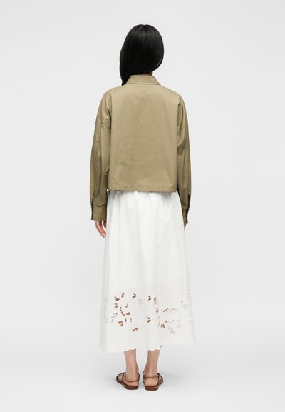 Γυναίκα που στέκεται με την πλάτη προς τα εμπρός, φοράει μπεζ μπουφάν, λευκή μίντι φούστα με κομμένο λουλουδάτο σχέδιο και καφέ σανδάλια σε λευκό φόντο.