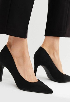 Chaussures à talons hauts en suède noir avec un bout pointu et un décolleté en V, dotées d'un talon aiguille et d'une texture lisse, associées à un pantalon noir.