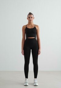 Must have mustavärvi sportlik crop top õhukeste rihmadega, kombineeritud kõrge vöökohtaga mustade retuusidega. Mudel kannab valgeid spordijalatsid ja seisab neutraalses taustas.