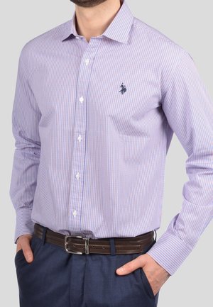 Man draagt een lichtpaarse geruite blouse met knopen en een logo op de borst, donkerblauwe broek en een bruine leren riem, handen in de zakken.