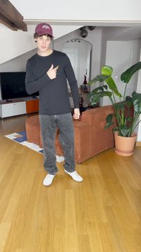 Černá košile s dlouhým rukávem, šedé džíny a bílé boty. Stojí v pokoji s dřevěnou podlahou, hnědou pohovkou a zelenými rostlinami v pozadí.