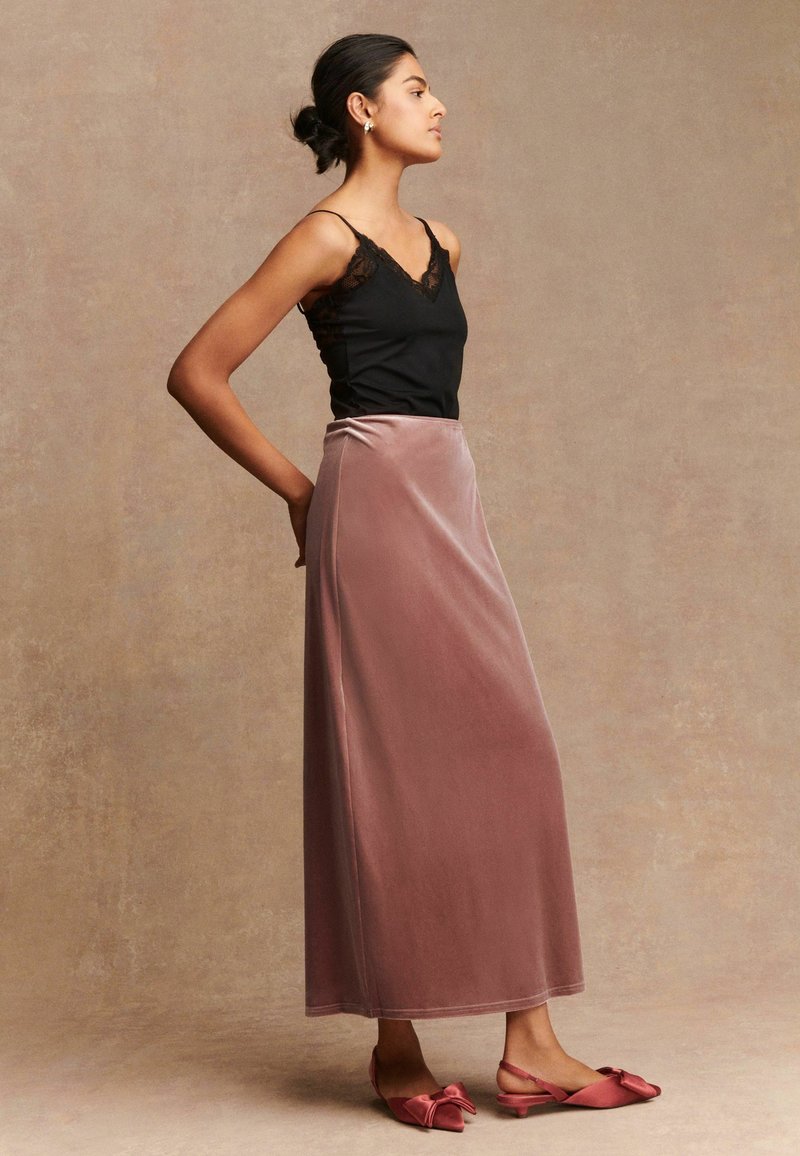 Vrouw in een zwarte camisole met kantafwerking, een lichtroze fluwelen maxi-rok en bijpassende lichtroze slingback hakken met strik, staand in zijaanzicht tegen een beige muur.