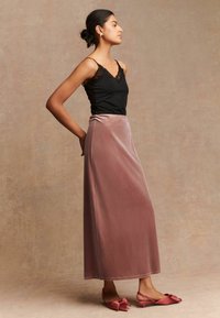 Schwarzes Spitzen-Camisole kombiniert mit einem langen, weichen rosa Samtrock und rosa Ballerinas mit Schleifenakzenten, stehend vor einem neutralen Hintergrund.