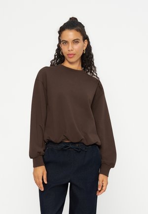 Donna con capelli ricci raccolti in una crocchia, che indossa orecchini a cerchio dorati, una felpa marrone a maniche lunghe e pantaloni blu scuro con coulisse, su sfondo bianco.