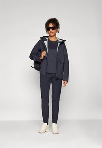 Tamsiai mėlyna lauko striukė su šviesiai pilkomis detalėmis, derinama su atitinkančia marškinėlių ir kelnių komplektu. Komplektą papildo juodas kuprinė ir kreminės spalvos sportbačiai.