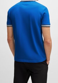 Mężczyzna w niebieskiej koszulce polo z krótkim rękawem, z białymi i czarnymi paskami na mankietach, stoi z rękami w kieszeniach czarnych spodni.